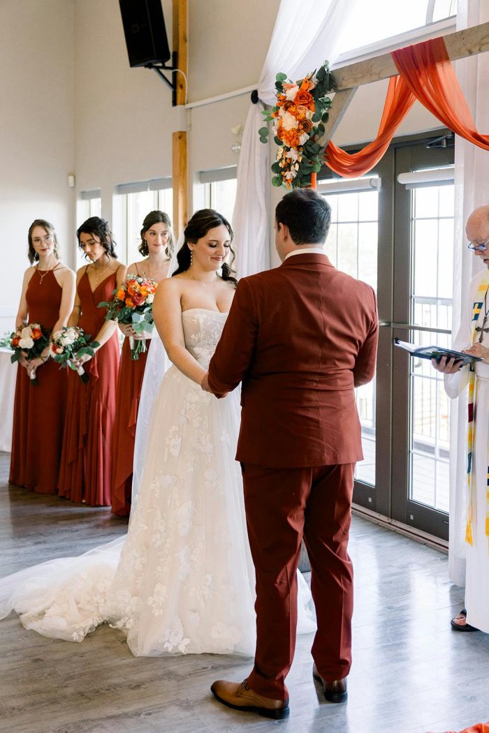 Bride in the Laila by Maggie Sottero lace wedding dress stands at the altar with her partner as bridesmaids in rust gowns look on
