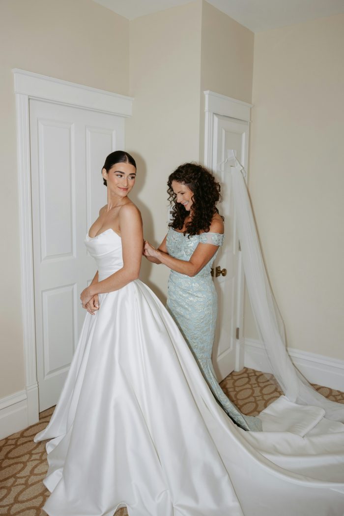 Bride wearing the Maureen by Maggie Sottero wedding dress while a bridesmaid helps adjust the gown during wedding day preparations