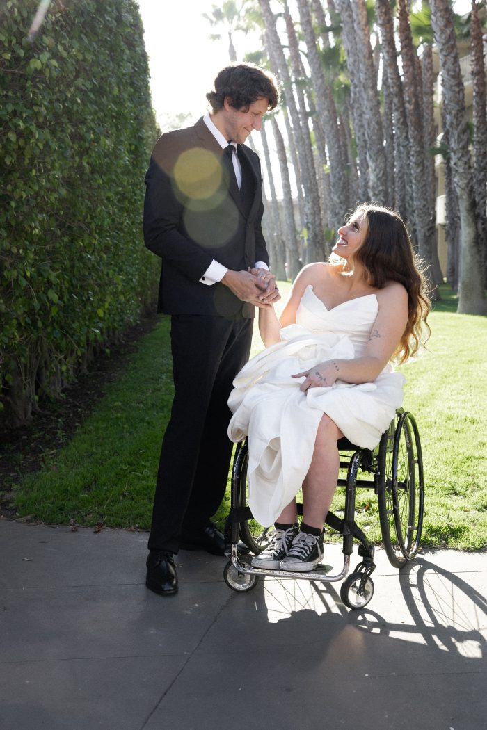Bride in a wheelchair wearing a simple wedding dress holding hands with her groom
