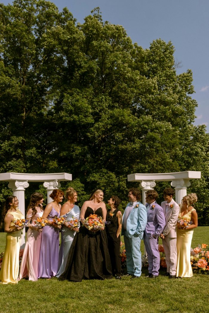Bride wearing the Derrick Marie by Maggie Sottero wedding dress poses with her wedding party in colorful pastel attire during an outdoor ceremony