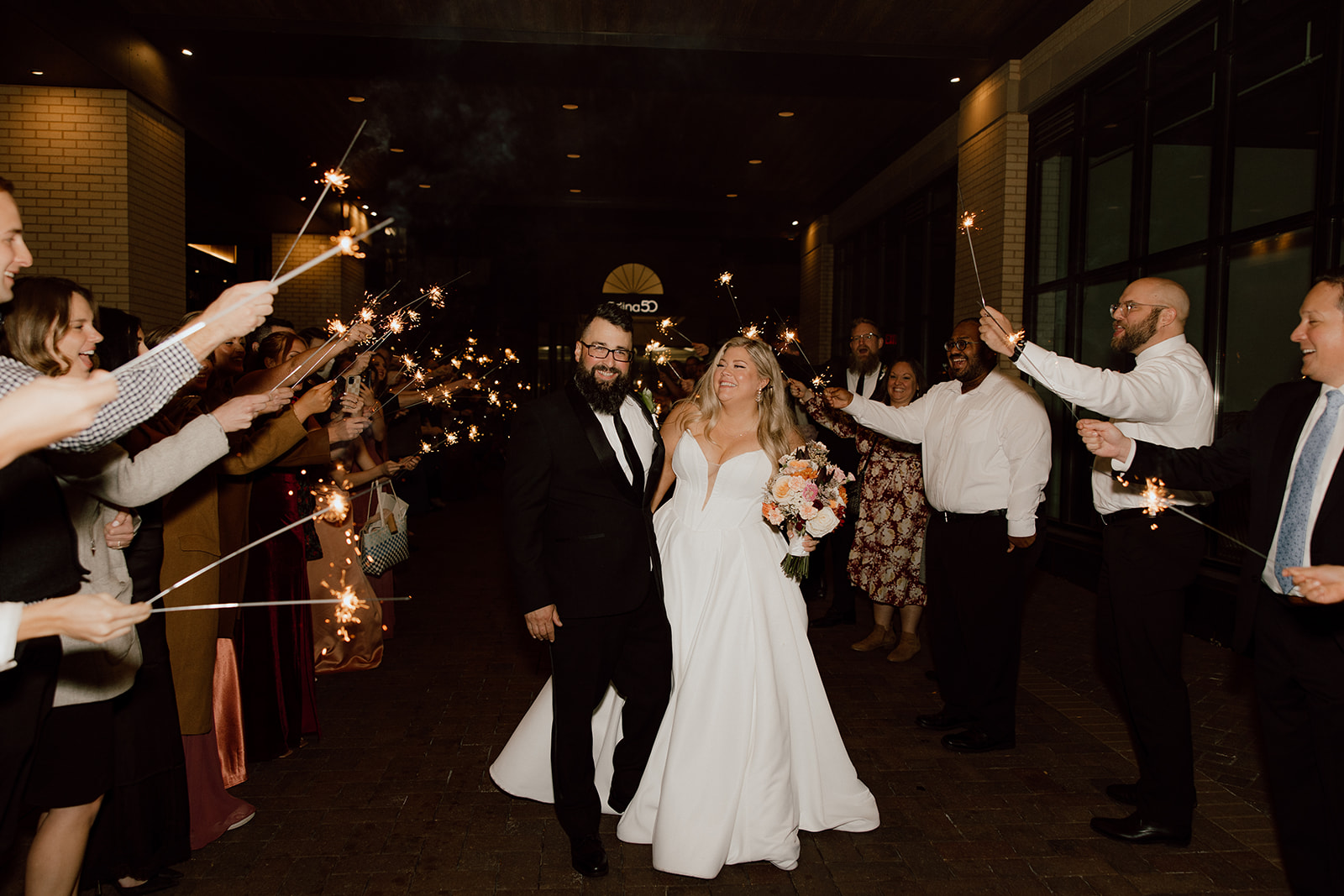 Bride wearing Derrick by Maggie Sottero walking down a hallway with her groom with guests holding sparklers