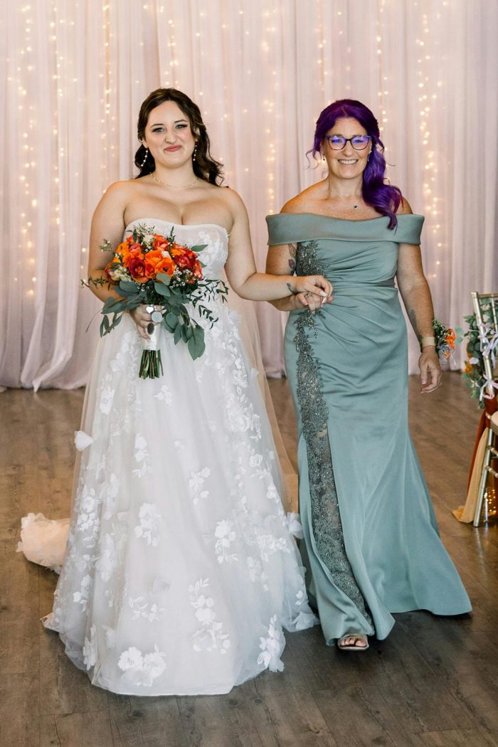 Bride wearing the Laila by Maggie Sottero wedding dress walks down the aisle holding a bouquet, accompanied by a bridesmaid in a sage green gown
