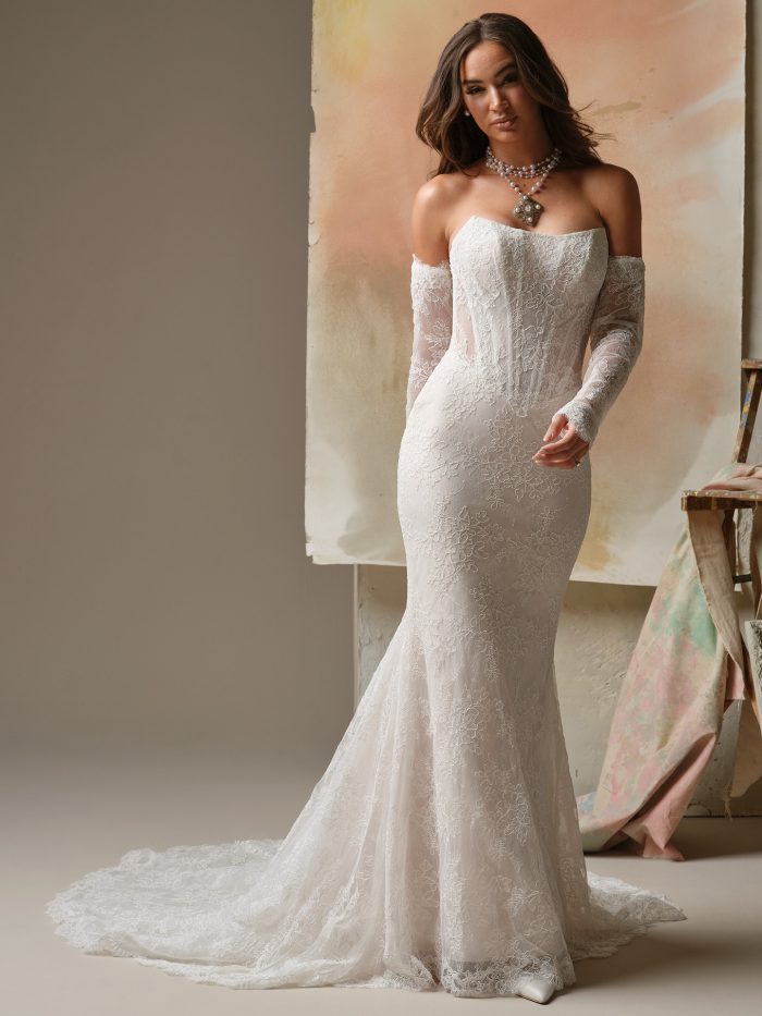 Bride wearing a romantic lace sheath wedding dress featuring a strapless neckline, exposed boning, and elegant off-the-shoulder lace sleeves. The gown’s fitted silhouette flows into a delicate train for a timeless and modern bridal look.