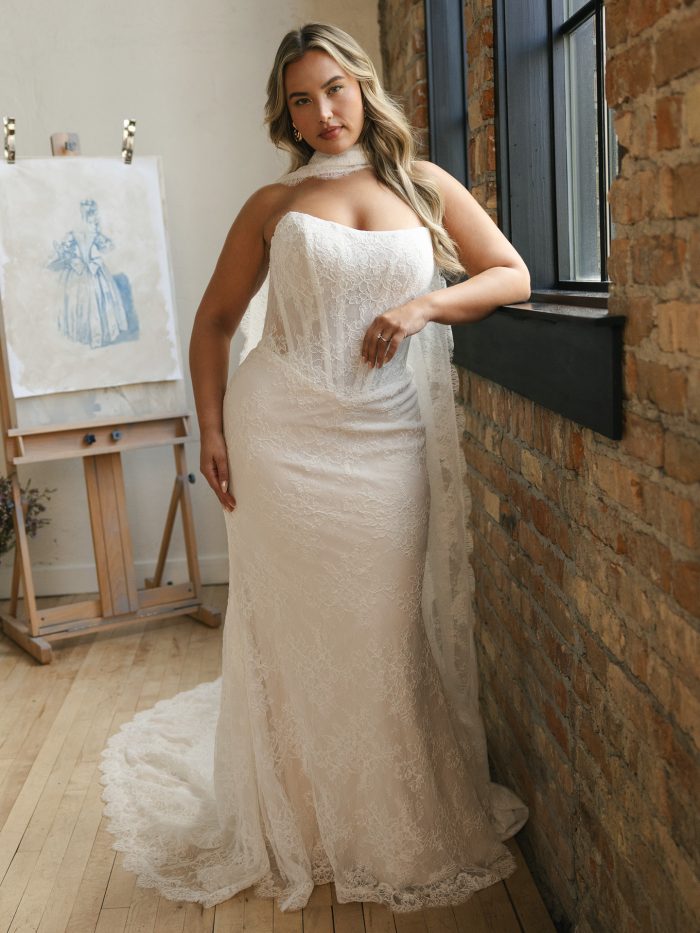 Curve bride wearing the Ariadne sheath wedding dress by Maggie Sottero, featuring a strapless neckline, soft floral lace, exposed semi-sheer corset detail, and a sweep train, styled with a delicate lace neck scarf.