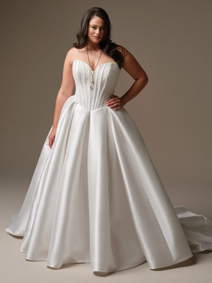 Curve model wearing Ashland by Maggie Sottero — strapless satin ballgown wedding dress with exposed boning, pleated skirt, and lace-embroidered bodice, styled with gold jewelry and side-swept curls.