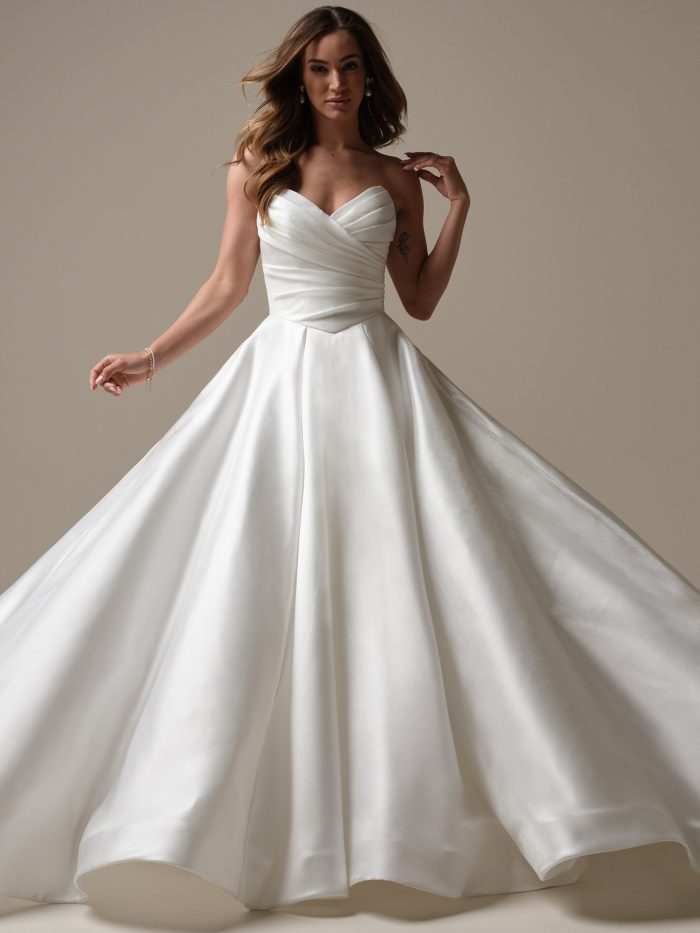 Aubrielle by Maggie Sottero — strapless sweetheart ballgown wedding dress featuring a ruched wrap-style bodice and full structured satin skirt, styled with loose waves in a neutral studio setting.