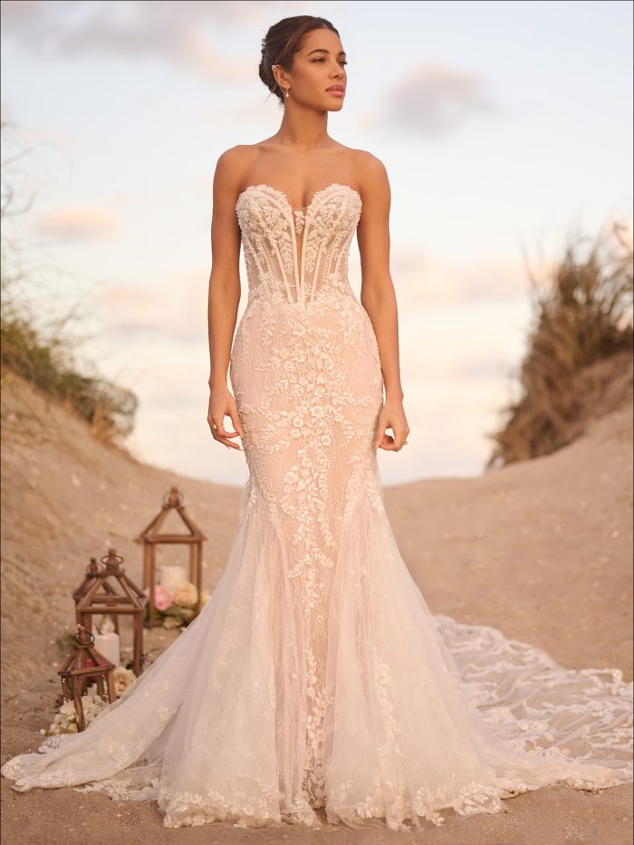 Model wearing the Benjie Royale by Maggie Sottero strapless fit-and-flare wedding dress with an illusion corset bodice, exposed boning, floral lace appliqués, and a soft lace train on a beach path.