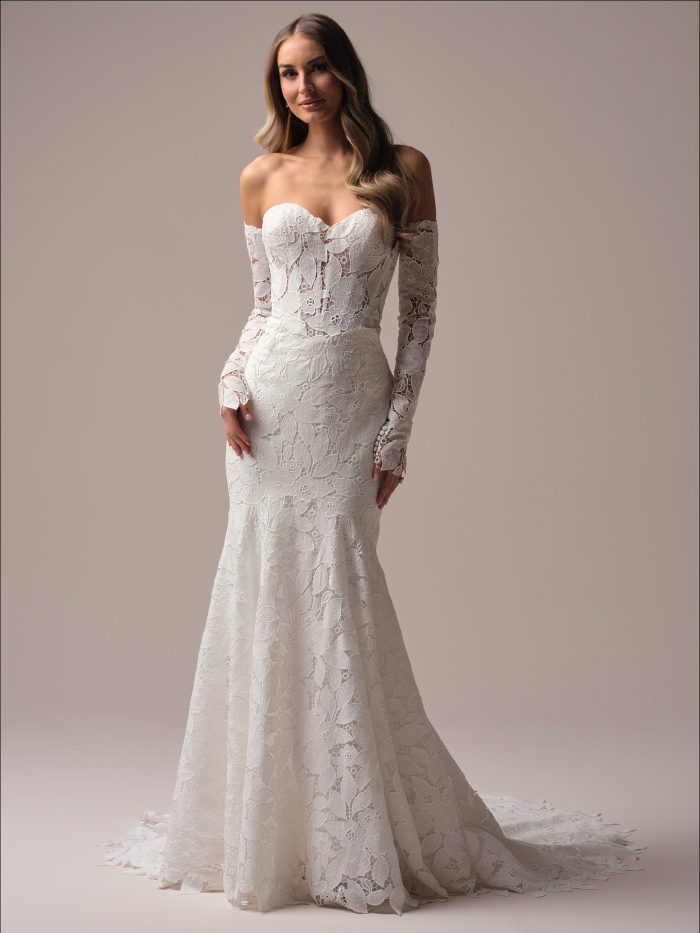 Bride wearing the Dakota fit-and-flare wedding dress featuring an off-the-shoulder sweetheart neckline, allover floral lace, sheer corset bodice, long lace sleeves, and a lace-trimmed train, photographed in a studio setting.