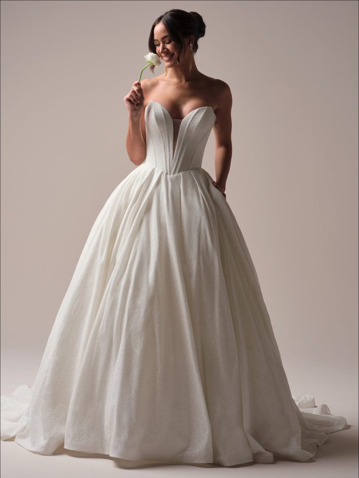 Bride wearing a strapless glitter tulle ball gown wedding dress with a deep illusion plunge neckline and structured bodice, smiling while holding a white flower.