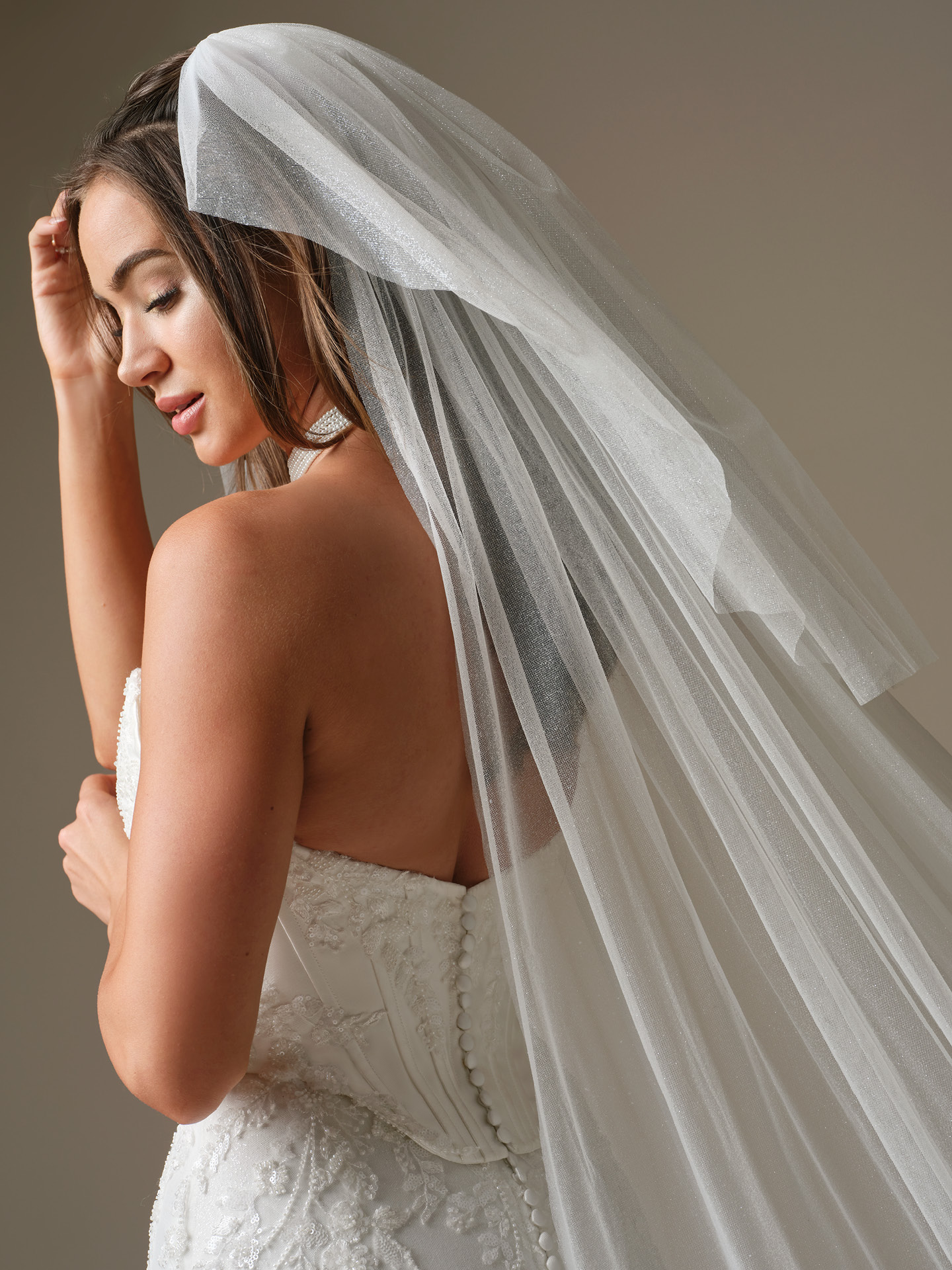 Bride wearing a sparkle tulle veil from the Maggie Sottero Essentials Edit, featuring a soft fingertip length, subtle shimmer throughout, and a light, airy drape over a strapless lace wedding gown with covered button detailing.