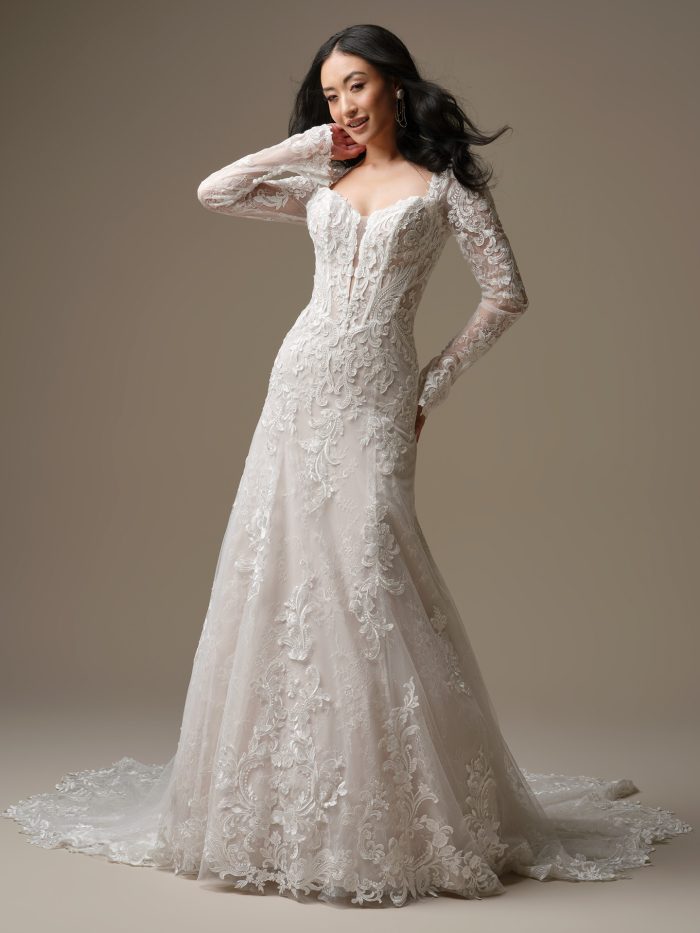 Bride wearing the Farrah A-line wedding dress by Maggie Sottero, featuring long illusion lace sleeves, a deep sweetheart neckline, exposed corsetry, and ornate embroidered lace over a soft tulle skirt and train.