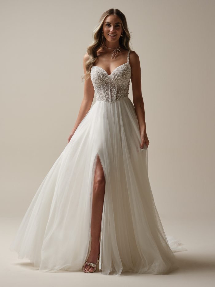 Bride wearing the Gretchen A-line wedding dress featuring a sheer pearl-beaded bodice with exposed boning, V-neckline, beaded spaghetti straps, and a soft tulle skirt with a side slit.