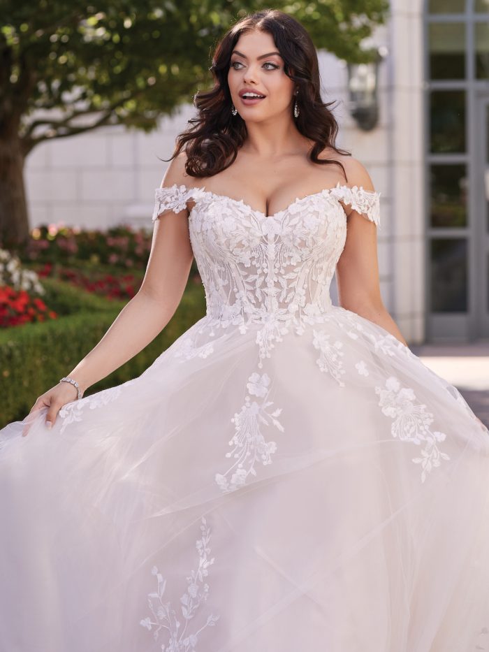 Bride in off-the-shoulder ball gown wedding dress with sweetheart neckline, floral lace appliqués, and layered tulle skirt, styled in an outdoor garden setting