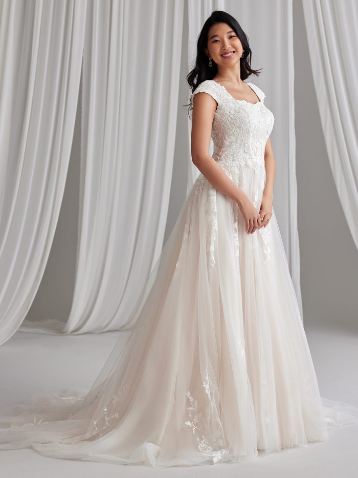 Bride wearing Maggie Sottero Harlem Leigh, an A-line modest wedding dress featuring a square neckline, cap sleeves, lace motifs with sequins over tulle, and a flowing skirt with train.