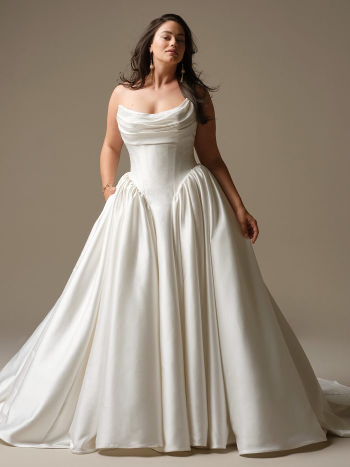 Model wearing Hudson by Maggie Sottero, a strapless Cartago satin ball gown wedding dress with dramatic draping, full skirt, and pockets.