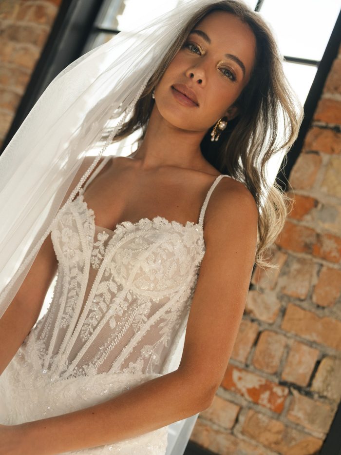Close-up of a bride in a beaded lace sheath wedding dress with a sheer corset bodice and delicate straps. She poses by a sunlit brick wall, highlighting the gown’s shimmering details and romantic, contemporary design.