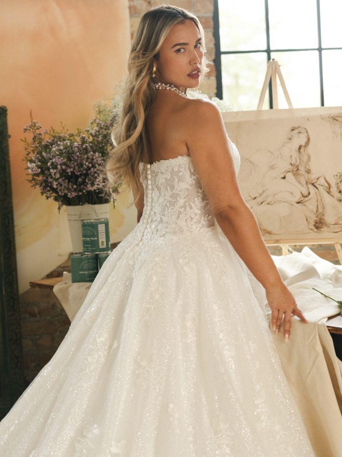 Back view of a strapless lace ballgown wedding dress with shimmering tulle layers and covered buttons. The bride’s long waves and pearl necklace complement the gown’s floral lace details and elegant, timeless silhouette.