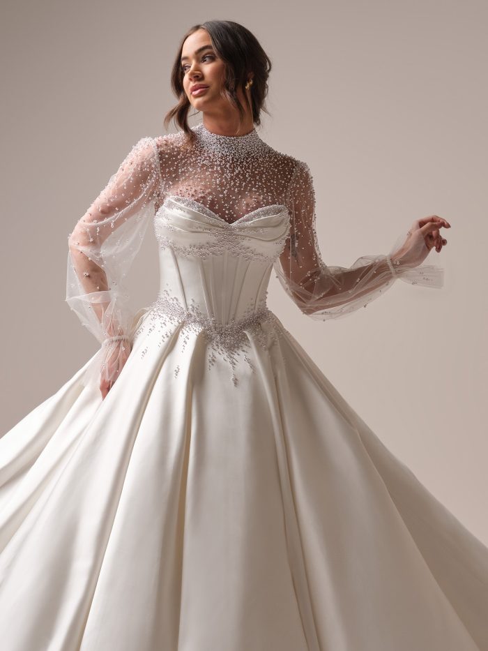 Bride wearing a strapless satin ball gown wedding dress styled with a sheer pearl-beaded illusion jacket featuring long sleeves and high neckline, posing elegantly in soft lighting.