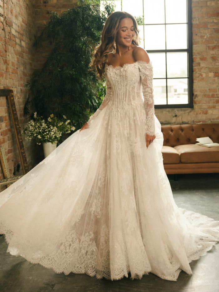 Bride wearing the Kitaro ballgown wedding dress by Maggie Sottero, featuring off-the-shoulder lace sleeves, exposed corsetry, 3-D floral appliqués, and a full lace skirt in a romantic loft setting.