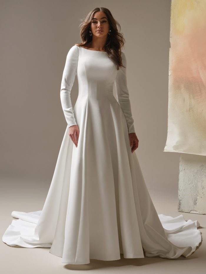 Lawrence wedding dress featuring a smooth long-sleeve bodice, a bateau neckline, a fitted waist, and a full A-line skirt that extends into a soft sweeping train. Model stands facing forward in a minimal studio setting.