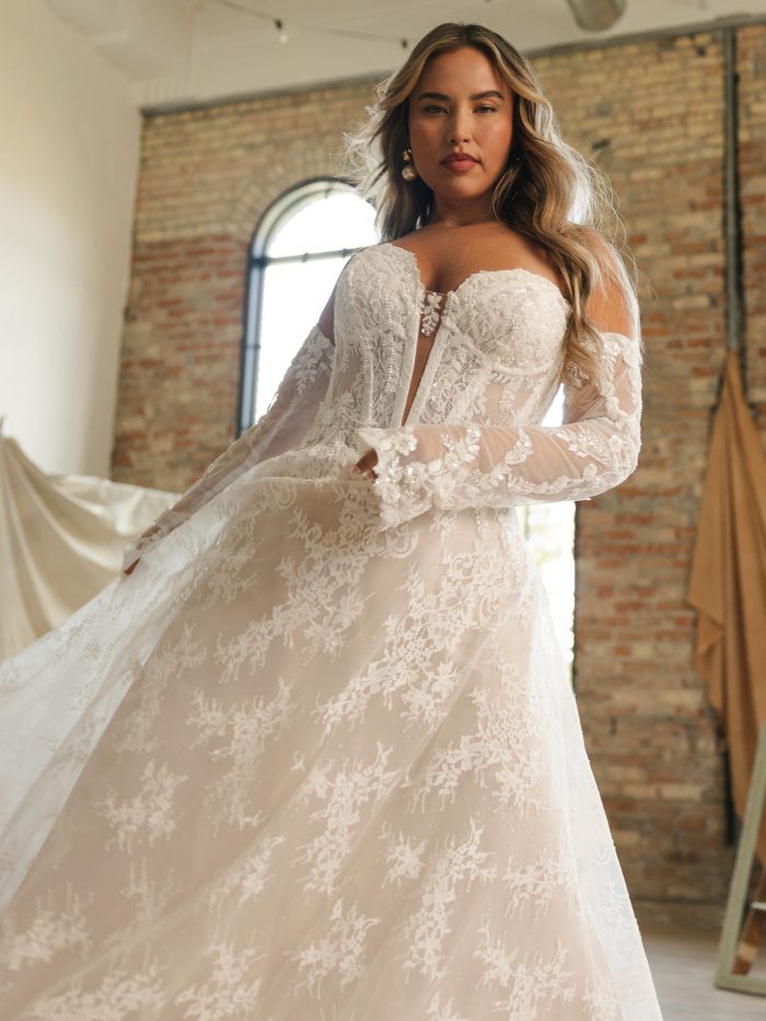 Lucca A-line wedding dress with off the shoulder illusion sleeves, shimmering lace embroidery, a deep illusion bodice, and a flowing lace trimmed skirt captured in natural light.