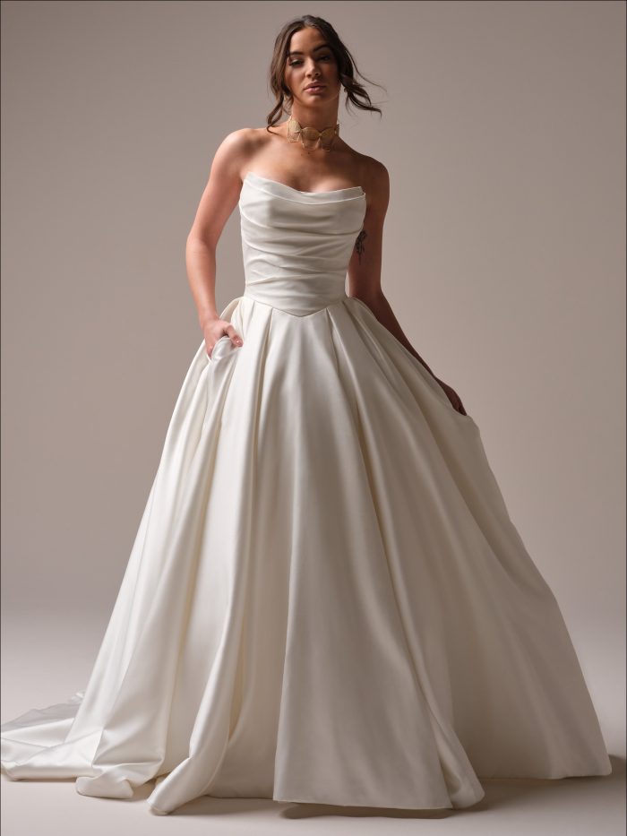 Bride wearing the Maureen ball gown wedding dress featuring a draped satin bodice, strapless neckline, and full pleated skirt with hidden pockets.