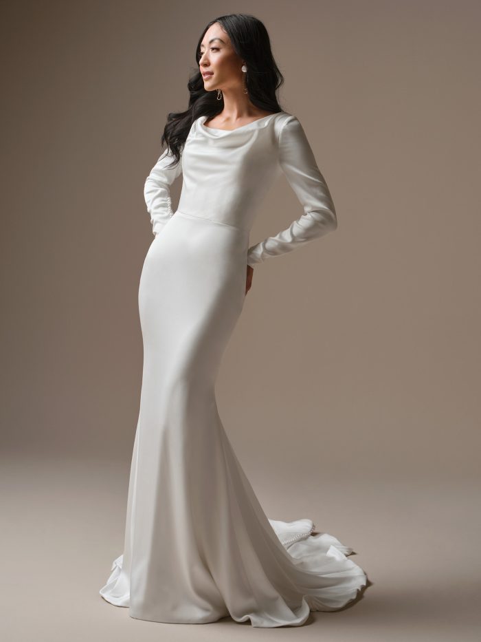Bride wearing Melinda by Maggie Sottero, a long-sleeve satin sheath wedding dress featuring a soft draped neckline, fitted silhouette, and an elegant chapel-length train.
