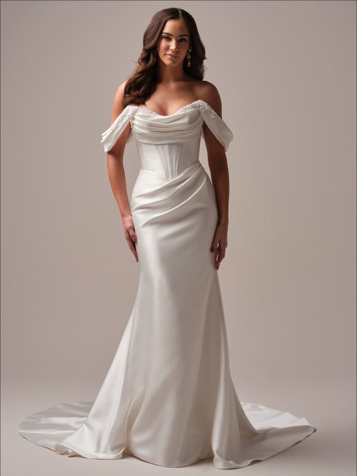 : Bride wearing the Melissa fit-and-flare wedding dress featuring luxurious Cartago satin, an asymmetrically draped bodice with pearl and bead accents, and off-the-shoulder straps for a romantic, timeless look.