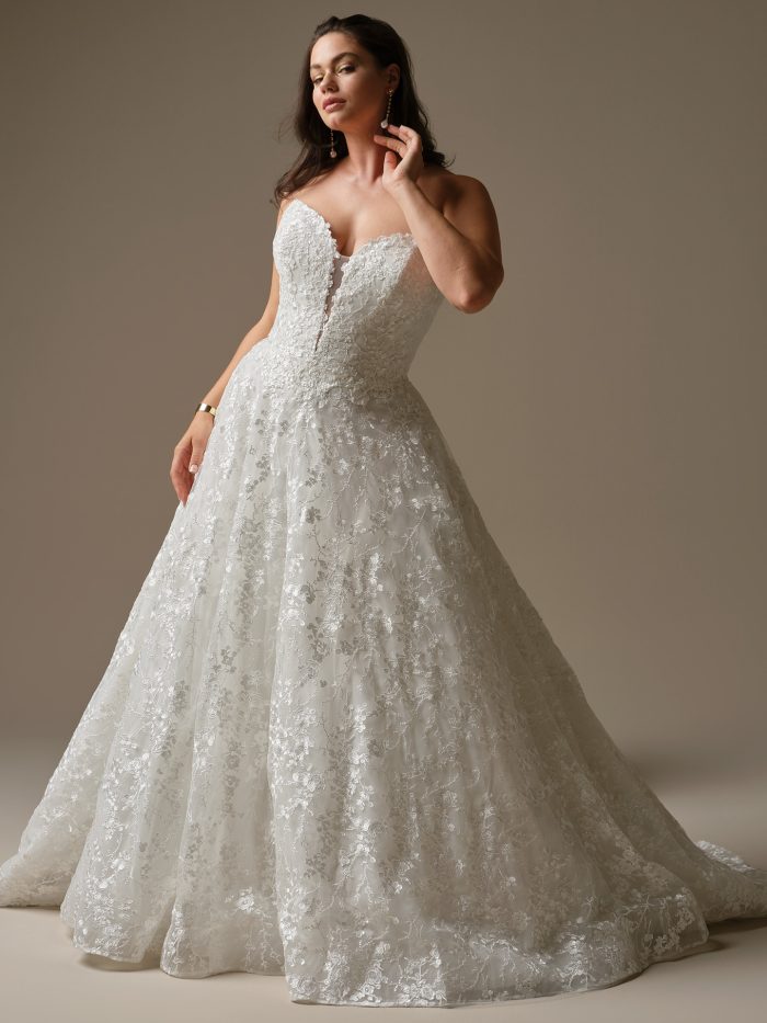 Model wearing Memphis by Maggie Sottero, a strapless ball gown wedding dress with sequined allover lace, plunging illusion V-neckline, and glitter tulle skirt.