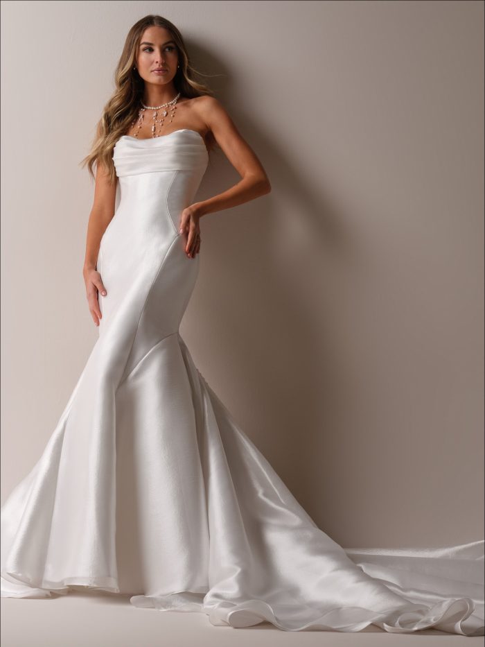 Odette Marie by Maggie Sottero — strapless satin fit-and-flare wedding dress with a softly draped bodice, sleek contour seams, and a dramatic flared skirt and train, styled with cascading pearl jewelry in a neutral studio setting.
