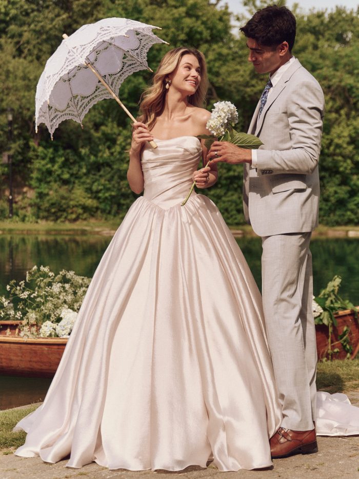 Bride in the Maggie Sottero Paris ballgown wedding dress with a strapless satin bodice and full skirt, holding a lace parasol beside a groom in a light suit by a lakeside setting with hydrangeas.