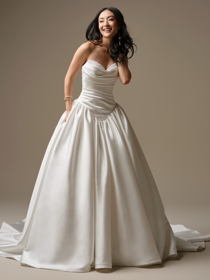 A model wearing the Maggie Sottero Pasadena wedding dress, featuring a draped Cartago satin bodice with an exaggerated basque waist, a pearl-accented sweetheart neckline and a full satin ball gown skirt with pockets.