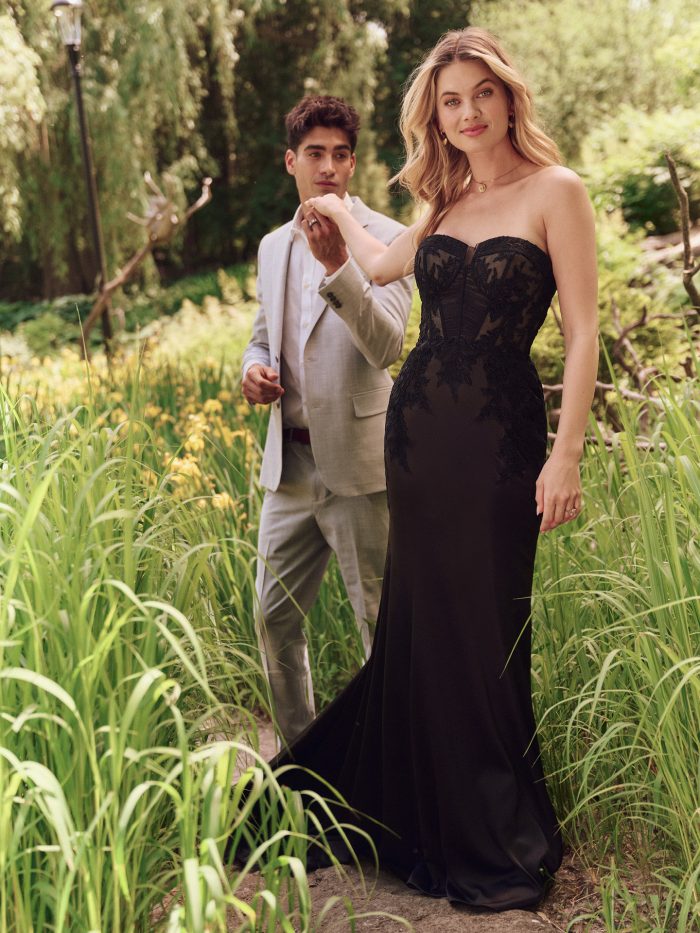 Bride wearing Persephone by Maggie Sottero, a dramatic black illusion lace sheath wedding dress with strapless neckline and corset-style bodice, walking outdoors with groom.