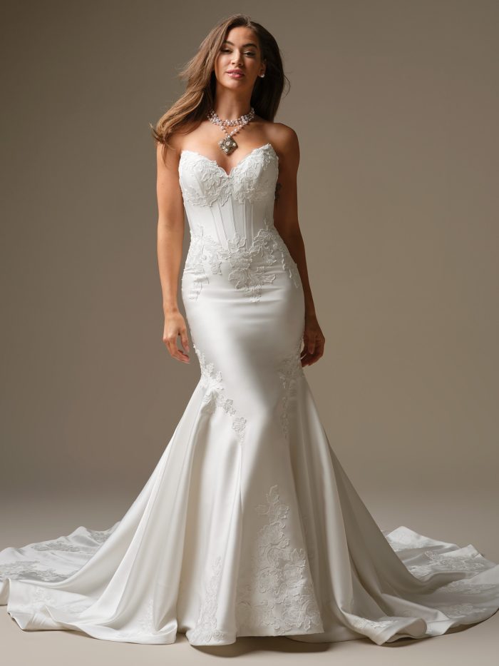 A model wearing the Maggie Sottero Serafina Lane wedding dress, featuring a strapless sweetheart neckline, corset bodice with lace motifs and a sleek fit-and-flare satin skirt with floral appliqués.