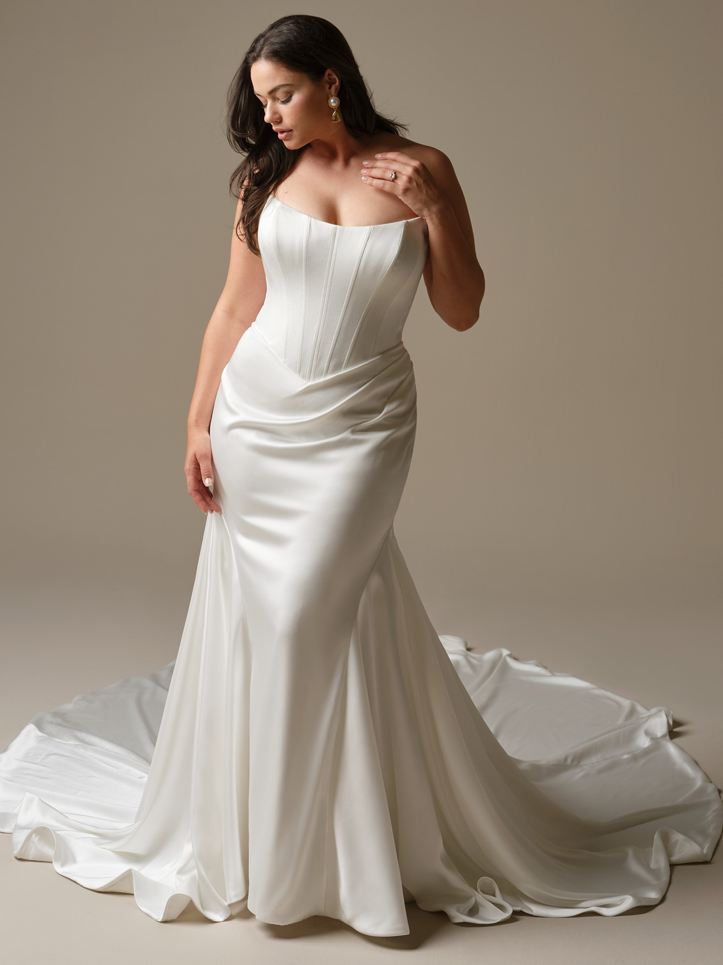 Bride wearing Sinclaire by Maggie Sottero, a strapless satin fit-and-flare wedding dress featuring a structured corset bodice, draped waist detail, and a sleek skirt that flares into a soft sweeping train.