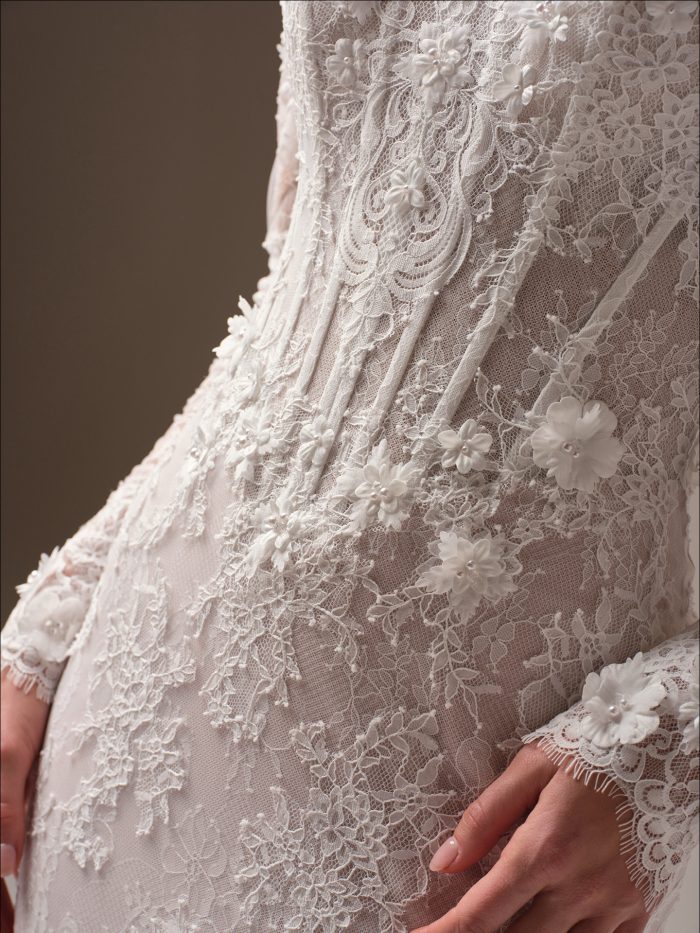 Close-up of the Takera bridal gown bodice highlighting exposed boning, Chantilly lace with 3-D floral appliqués accented with pearls, and scalloped lace sleeve cuff detail.