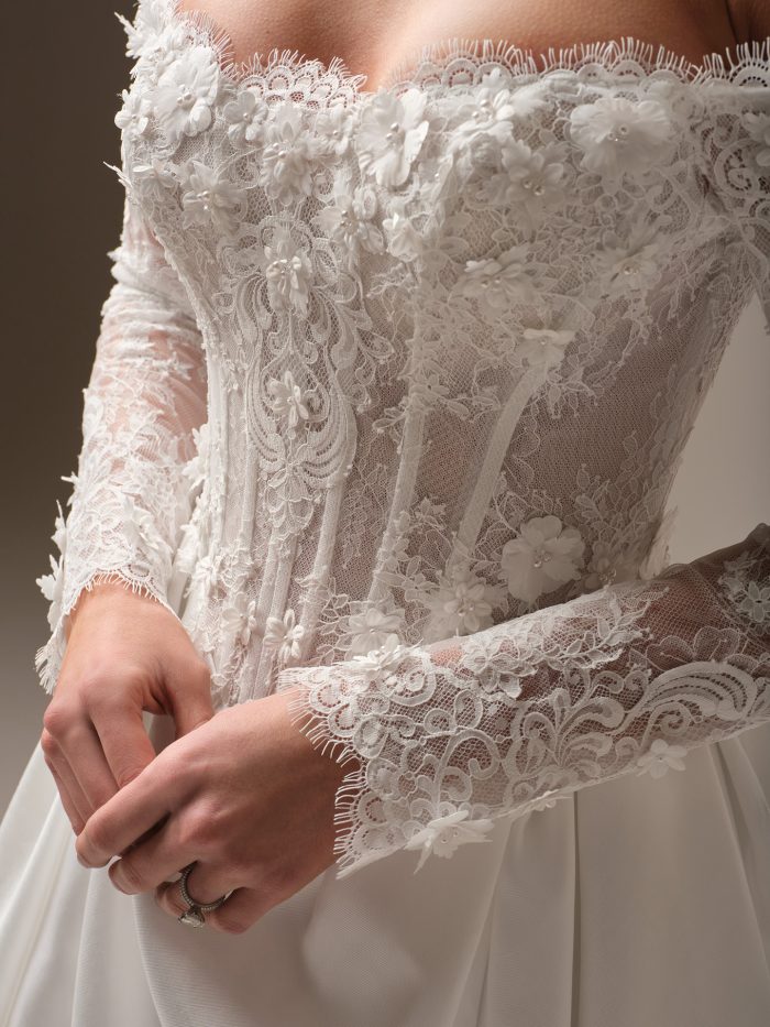 Takera Lane by Maggie Sottero — close-up of an off-the-shoulder corset bodice with illusion lace sleeves and 3-D floral appliqués, paired with a soft satin A-line skirt in a studio setting.