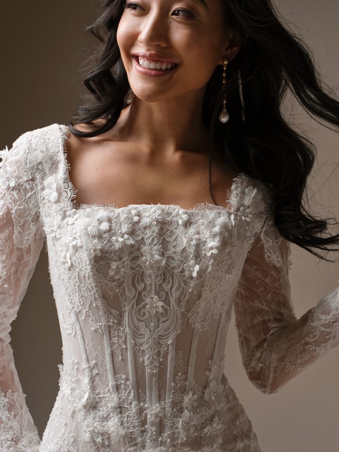 Close-up of the Takera Leigh wedding gown by Maggie Sottero, showcasing the square neckline, long illusion lace sleeves, exposed corset bodice, and intricate 3-D floral lace appliqués.