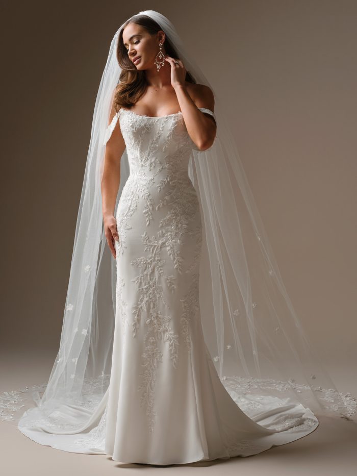 Bride wearing the Maggie Sottero Thera sheath wedding dress featuring a strapless neckline, fitted silhouette with floral lace appliqués, and chapel-length train. Styled with a long embroidered veil.