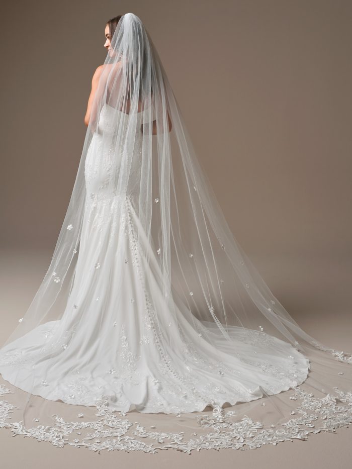 Bride wearing a cathedral-length veil adorned with delicate floral lace appliqués and a scalloped lace edge. The sheer tulle veil flows over a fitted wedding dress, creating a romantic and timeless bridal look from the back.