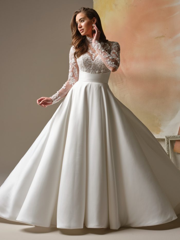Trinity by Maggie Sottero — high-neck long-sleeve A-line wedding dress featuring an illusion lace bodice with floral embroidery, a defined waist, and a full satin skirt, styled in a soft studio setting.