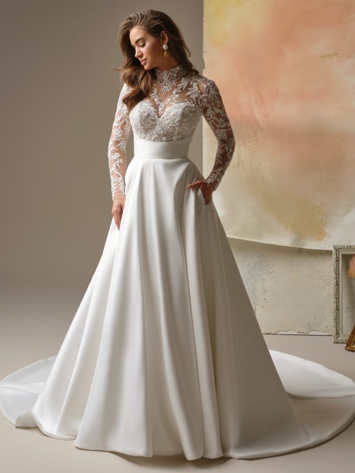 Trinity wedding dress featuring a Lyric Mikado A-line skirt with pockets, an illusion beaded and sequined lace bodice over a sweetheart base, a mock illusion lace turtleneck, long fitted illusion lace sleeves, an empire waistband with topstitch detail, and covered buttons trailing to the hem. Model stands in a softly lit studio setting.