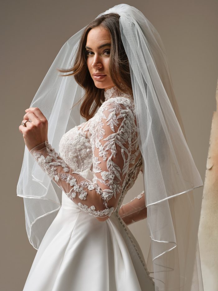 Close-up view of the Trinity wedding dress showing the mock illusion lace turtleneck, beaded and sequined lace bodice, fitted illusion sleeves with lace detailing, the empire waistband, and covered buttons along the back. Model wears a soft tulle veil.