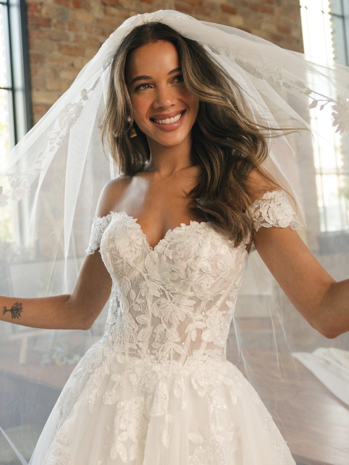 Verina ballgown wedding dress featuring off the shoulder floral lace, a semi sheer lace bodice, and a soft cathedral veil captured in natural window light.