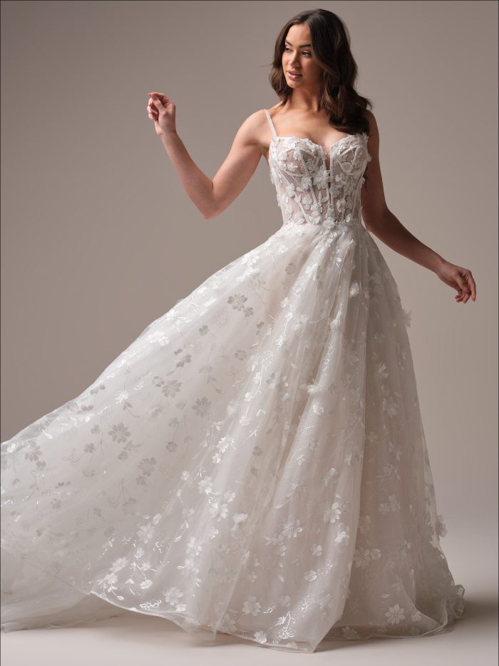 Bride wearing the Maggie Sottero Wallis ballgown wedding dress featuring a sheer corset bodice with delicate spaghetti straps, 3D floral appliqués, and a shimmering layered skirt for a romantic textured bridal look.