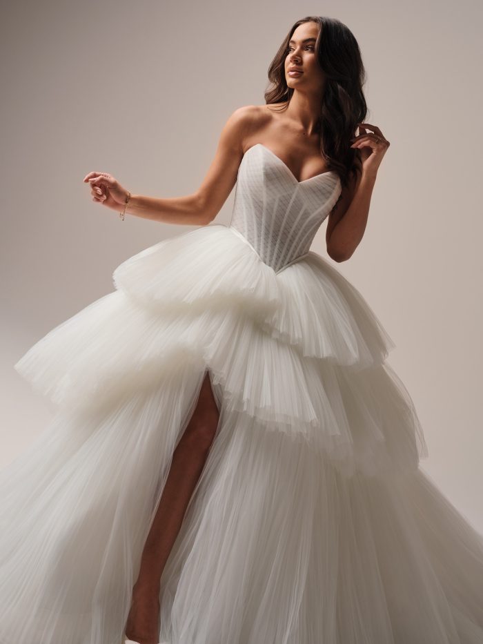 Bride wearing a strapless tiered tulle ball gown wedding dress with a structured corset bodice and high slit, posing gracefully in soft studio lighting.