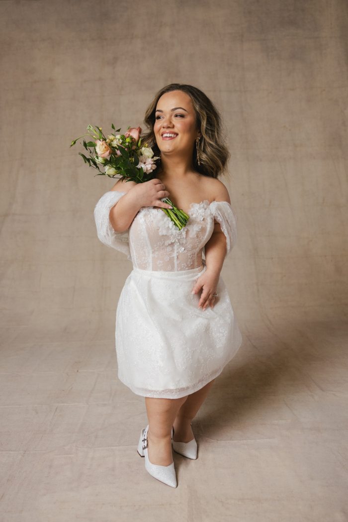 Little person bride wearing a sparkly mini wedding dress with off-the-shoulder cap sleeves holding a flower bouquet on her shoulder