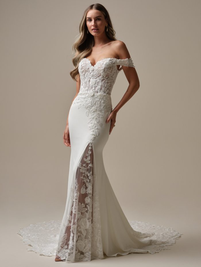 Model wearing the Rebecca Ingram Adeline sheath wedding dress with an off-the-shoulder Chantilly lace bodice, lace illusion slit, and fitted crepe skirt.