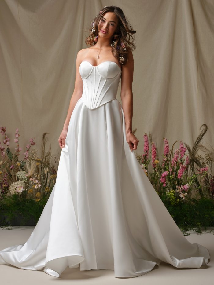 Model wearing the Rebecca Ingram Alexia A-line wedding dress featuring a satin bodice with exposed boning, sweetheart neckline, and flowing skirt with pockets.