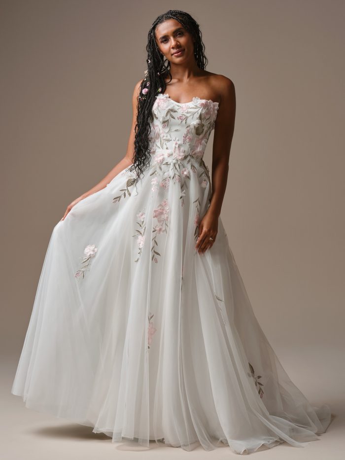 Bride wearing the Rebecca Ingram Blossom A-line wedding dress featuring a strapless sweetheart neckline, soft tulle skirt, and blush 3D floral appliqués with embroidered leafy details.